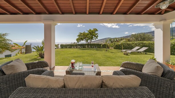 A covered patio with wicker furniture and a table set for two, overlooking a well-maintained lawn and distant hills.