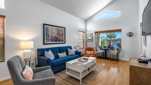 Un salon spacieux avec un canapé bleu, un fauteuil gris et une table basse. Un tableau orne le mur, une fenêtre est équipée de stores, et on peut admirer la vue sur l’extérieur.