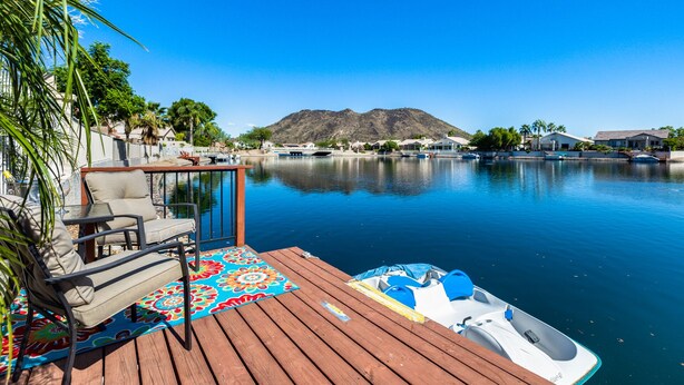 Une terrasse en bois agrémentée d’un tapis aux couleurs vives, d’un bateau, et offrant une vue dégagée sur un lac, avec des maisons et des montagnes en arrière-plan.