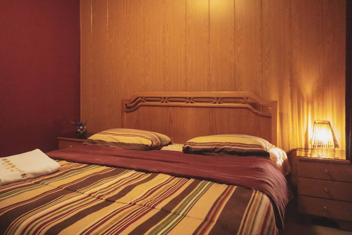 A hotel room with a wooden headboard, striped bedding, a bedside table with a lamp, and a folded towel on the bed.