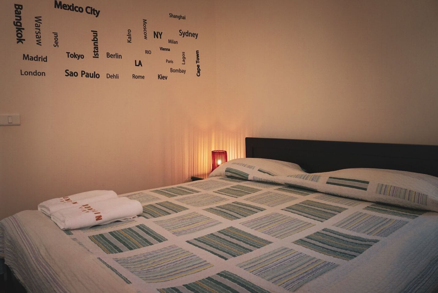 Ein ordentlich bezogenes Bett mit einem gemusterten Bettdecken und gefalteten Handtüchern, eine Nachttischlampe und eine Wand mit Städtenamen.