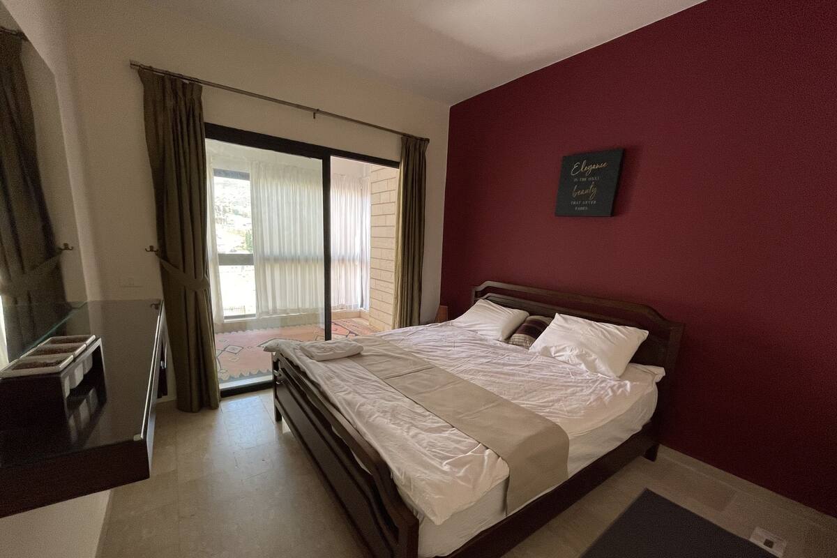 A bedroom with a large bed, a wooden headboard, a red accent wall, and a sliding glass door leading to a balcony.