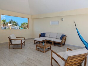 A spacious balcony with a wooden floor, a beige sofa with blue cushions, two wooden chairs, and a hammock.