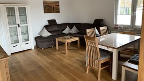 Uma sala de estar moderna com um sofá marrom escuro, uma mesa de centro de madeira e uma mesa de jantar com cadeiras. Há um armário branco com portas de vidro e um quadro na parede.