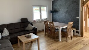 Uma sala de estar moderna com parede de pedra cinza escura, uma mesa de jantar de madeira com quatro cadeiras, um sofá marrom escuro com almofadas estampadas e uma pequena mesa de centro de madeira.