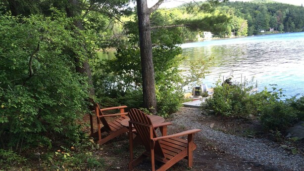 A wooden table and two chairs set up on a gravel path beside a lake, surrounded by dense greenery. - Honeymooners/Getaway Studio on Lovely Lake Iroquois near Burlington (Hinesburg)