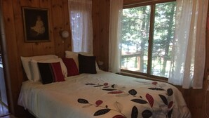 A wooden cabin bedroom with a bed, pillows, a framed picture on the wall, and a window with curtains. - Honeymooners/Getaway Studio on Lovely Lake Iroquois near Burlington (Hinesburg)
