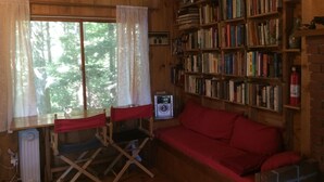 A cozy cabin interior with a wooden interior, a red couch, a bookshelf, and a window with curtains. - Honeymooners/Getaway Studio on Lovely Lake Iroquois near Burlington (Hinesburg)