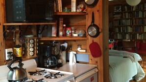 A compact kitchen with a stove, microwave, and various kitchen items. A bookshelf and a bed are visible in the background. - Honeymooners/Getaway Studio on Lovely Lake Iroquois near Burlington (Hinesburg)