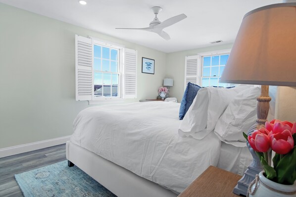 A bedroom with a bed, white walls, a ceiling fan, and a window with shutters. - *Oceanfront Beach Home* (Sandwich)