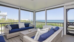 A covered outdoor seating area with wicker furniture and cushions, overlooking a beach and ocean. - *Oceanfront Beach Home* (Sandwich)