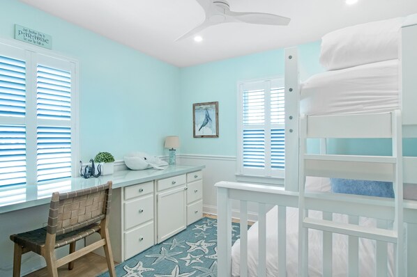 A room with a bunk bed, a desk with a chair, a ceiling fan, and a window with shutters. - *Oceanfront Beach Home* (Sandwich)