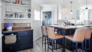 A modern kitchen with a central island, bar stools, and a bookshelf with decorative items. - *Oceanfront Beach Home* (Sandwich)