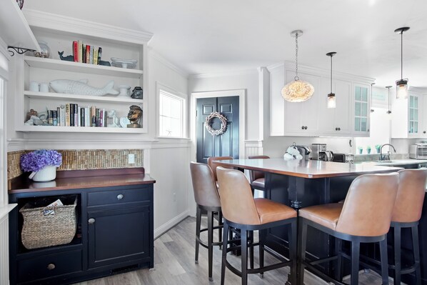 A modern kitchen with a central island, bar stools, and a bookshelf with decorative items. - *Oceanfront Beach Home* (Sandwich)