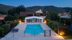 A swimming pool with a glass enclosure, surrounded by a fence, and a view of a residential area with houses and hills in the background. - Homerez - House in Saint-Géry + Pool (Saint-Géry)