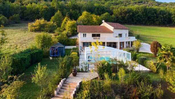 A two-story house with a large glass conservatory, surrounded by lush greenery and a hilly landscape. - Homerez - House in Saint-Géry + Pool (Saint-Géry)