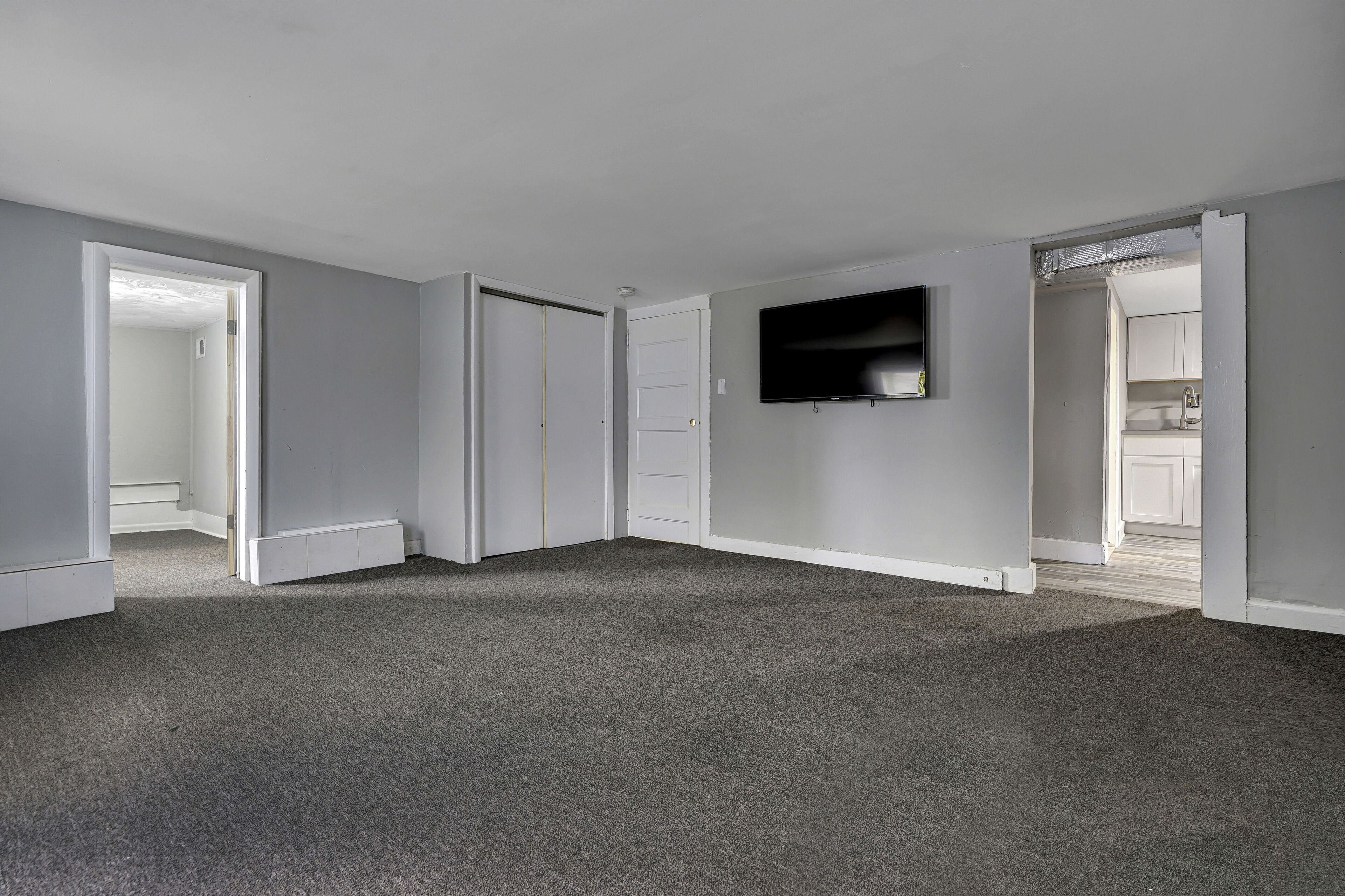 Una habitación con alfombra gris, paredes blancas, un televisor de pantalla plana montado en la pared y una puerta que da a una zona de cocina.