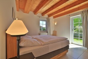 Une chambre à coucher avec un lit, une table de chevet, une lampe, une fenêtre et une vue sur la verdure à l’extérieur.