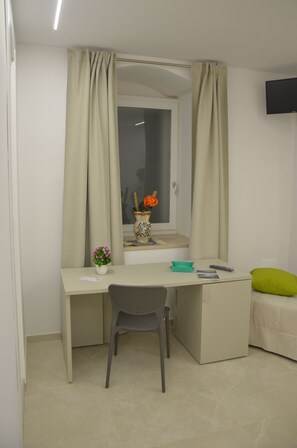 Une petite chambre à coucher, bien rangée, avec un lit simple, un bureau et une chaise, une fenêtre avec des rideaux et un téléviseur à écran plat fixé au mur.
