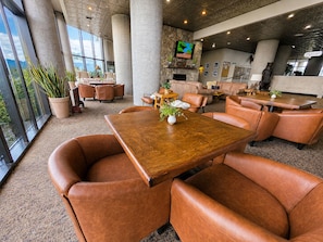 A spacious indoor area with a large wooden table surrounded by leather chairs, a stone wall with a television, and a view through large windows.
