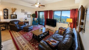 A living room with a sofa, a coffee table, a TV, and a large window with curtains. - Sugartop Resort 6th-Floor Corner Condo: Breathtaking Mountain Views - 2BR (Sugar Mountain)