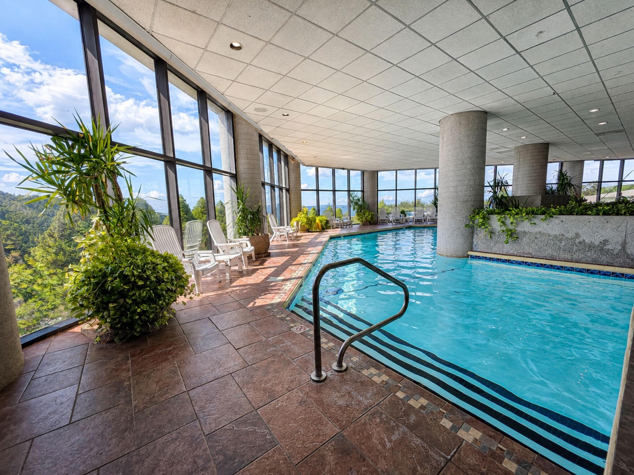 An indoor swimming pool with clear views of the surrounding landscape, surrounded by large windows and furnished with lounge chairs.
