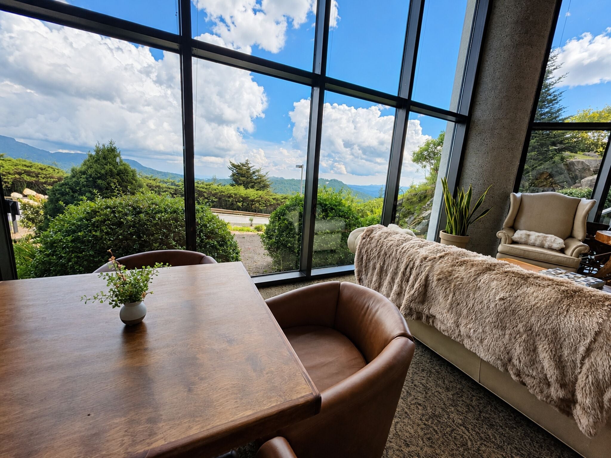 A room with a large window offering a view of a mountainous landscape. There is a wooden table with a vase of flowers and a brown leather chair.
