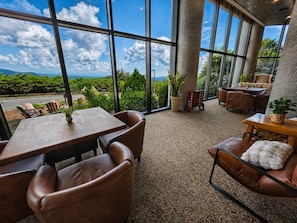A spacious indoor area with large windows offering a view of greenery and mountains. There are leather chairs, a wooden table, and a sofa with a fluffy pillow.