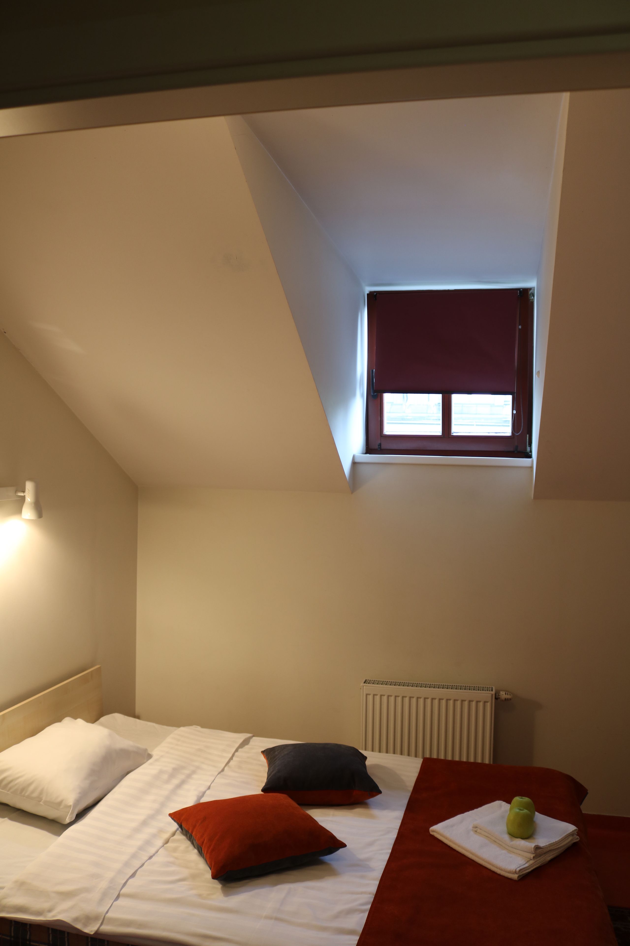 Une chambre avec un plafond en pente, une petite fenêtre avec un store rouge, un radiateur et un lit bien fait, agrémenté d’oreillers.