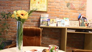 A table with a book, a muffin, and a cup of coffee, a vase with flowers, and a brick wall with a framed picture and a shelf with books and cards. - Apple Economy Hotel (Kaunas)