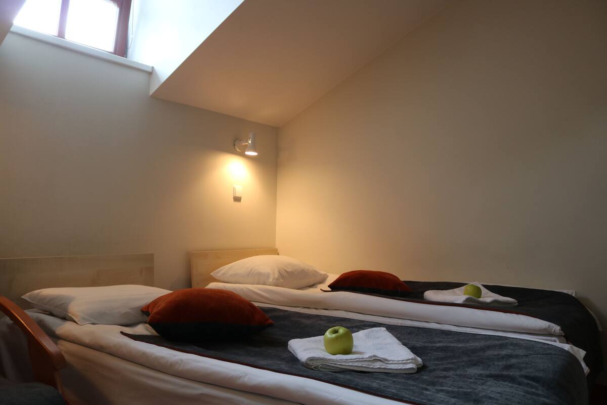 A hotel room with two beds, each with white and red pillows, a small apple on a towel, and a wall-mounted light.