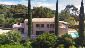 Une maison à deux étages avec une piscine, entourée de grands arbres et située dans un paysage vallonné.