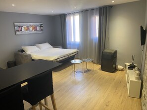 A modern hotel room with a bed, a desk, two side tables, a TV, and a large window with curtains.