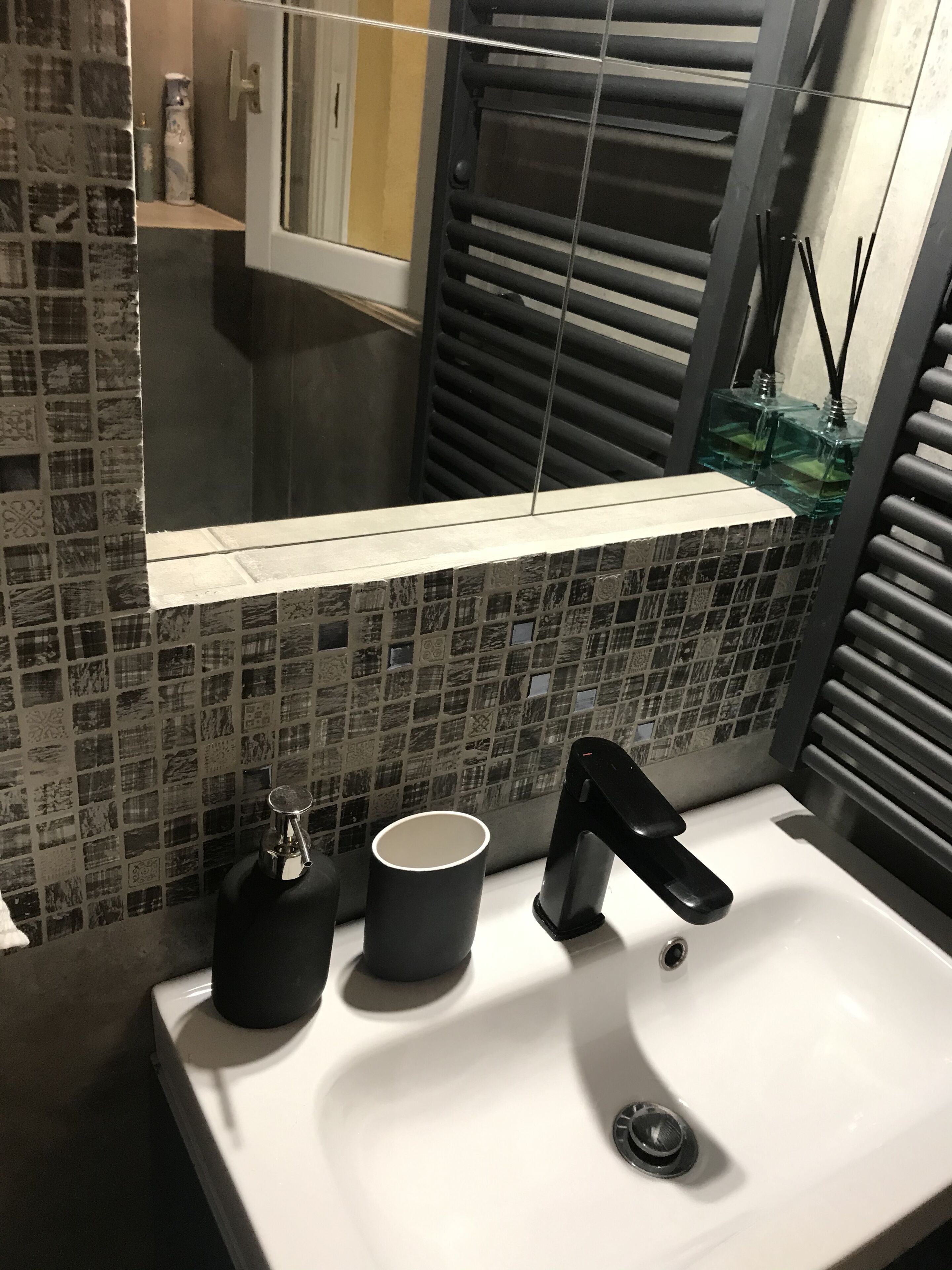 A bathroom with a white sink, a black faucet, and a mirror above it. There is a window with a view of a room with a bathtub and a towel rack.