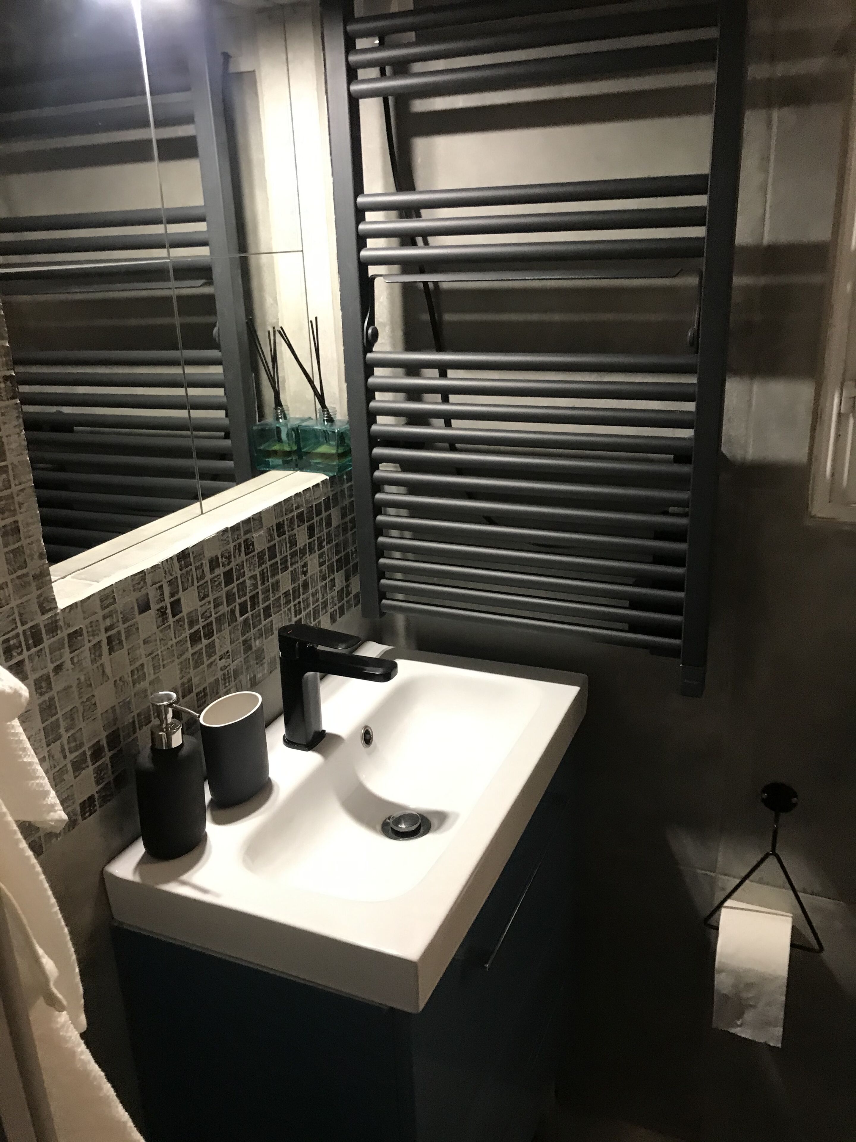 A bathroom with a white sink, black faucet, and a mirror above it. There is a window with blinds, a towel rack, and a bottle of hand soap on the sink.