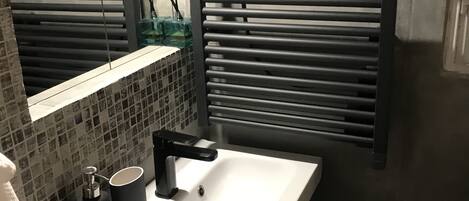 A bathroom with a white sink, black faucet, and a mirror above it. There is a window with blinds, a towel rack, and a bottle of hand soap on the sink.