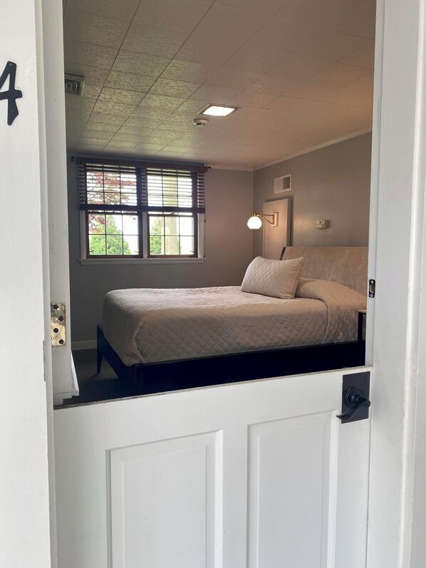 Une chambre à coucher avec un lit, une fenêtre avec des stores, un luminaire fixé au mur et une porte sur laquelle est inscrit le numéro « 4 ».