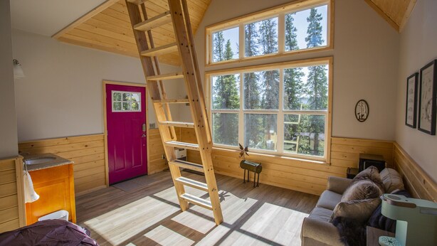 A cozy cabin interior with a wooden ladder, a red door, and a large window overlooking trees. - Alpenglow Ridge Fireweed (Healy)