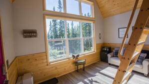 A cabin with a large window offering a forest view, a wooden ladder, and a cozy seating area. - Alpenglow Ridge Fireweed (Healy)