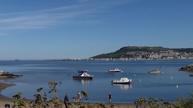 Uma vila costeira com barcos na água, uma praia de areia e uma colina com edifícios ao fundo.