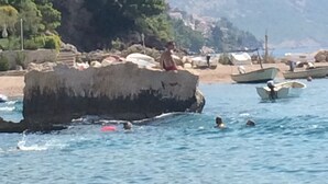 Paisaje costero con terreno rocoso, barcos y una persona sentada en una roca junto al agua.