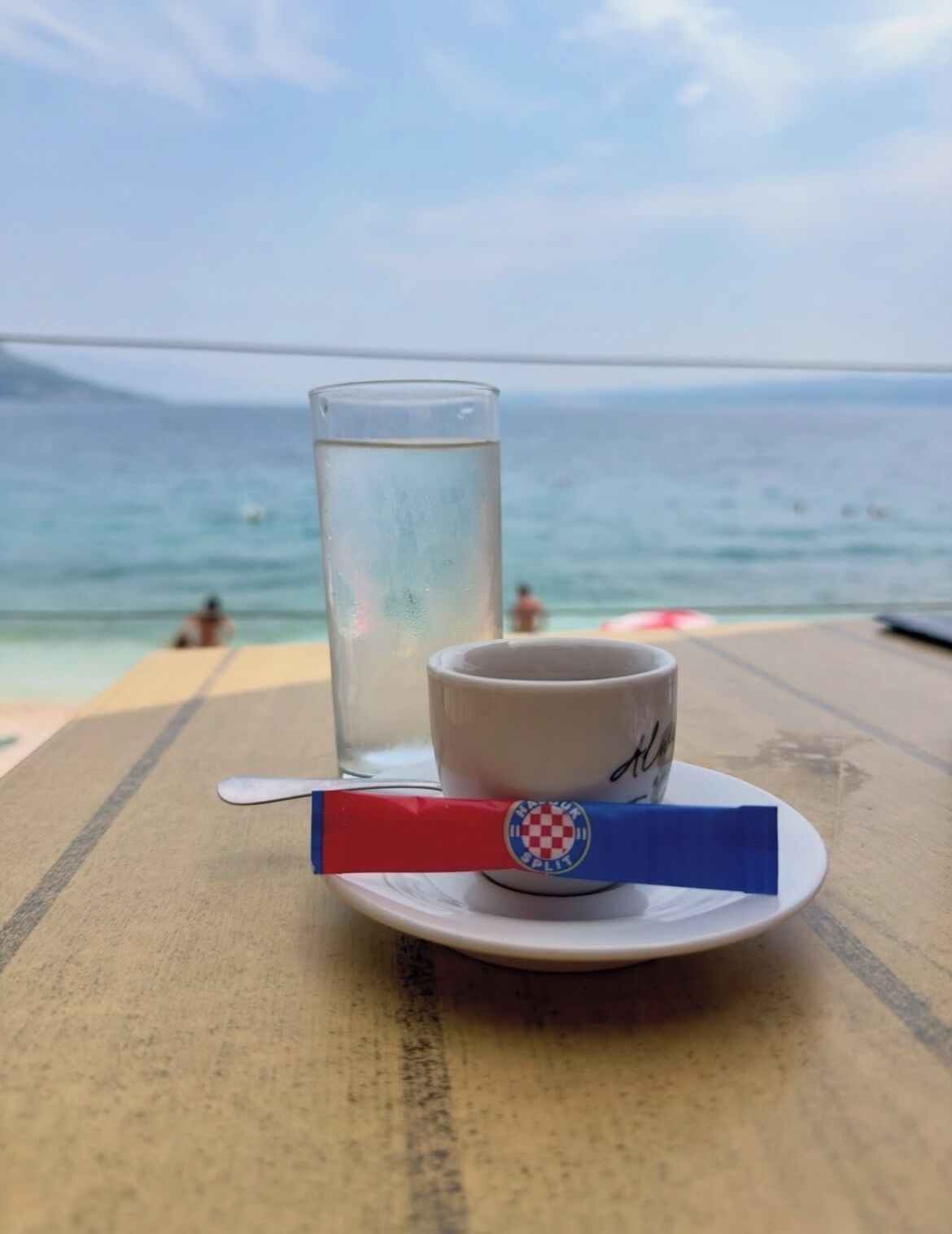 A table with a glass of water and a mug on a plate, overlooking a beach with people in the water.