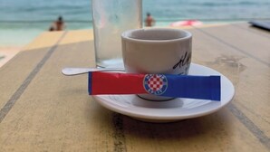 Una mesa con un vaso de agua y una taza sobre un plato, con vista a una playa donde hay gente en el agua.