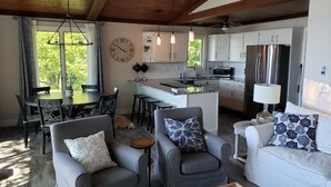 Una cocina moderna con armarios blancos, electrodomésticos de acero inoxidable e iluminación colgante. Hay un comedor con una mesa redonda y sillas, y una sala de estar con sofá y sillones.