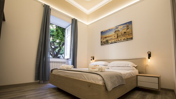 A modern bedroom with a large bed, a nightstand, a painting of a historical building, and a window with curtains. - Sammartino suite (Palermo)