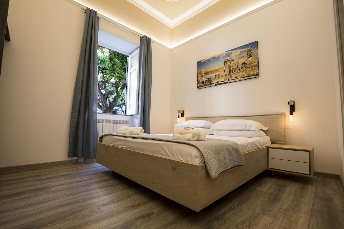 Ein modernes Schlafzimmer mit einem großen Bett, einem Nachttisch, einem Gemälde eines historischen Gebäudes und einem Fenster mit Vorhängen.
