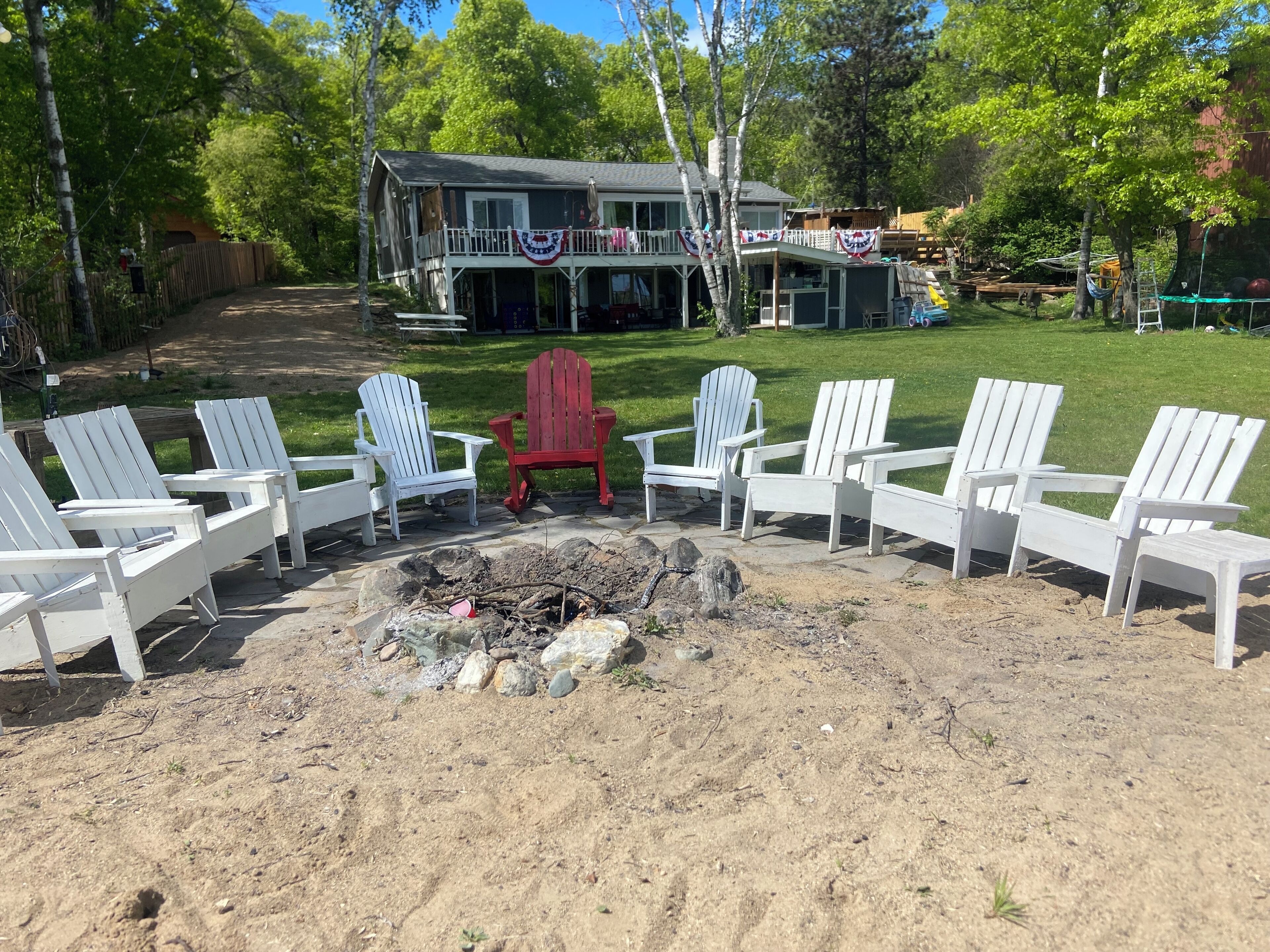 A backyard with a fire pit surrounded by Adirondack chairs, a wooden deck, and a house with a porch.