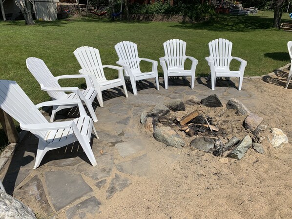 A fire pit surrounded by white Adirondack chairs in a stone-paved area.