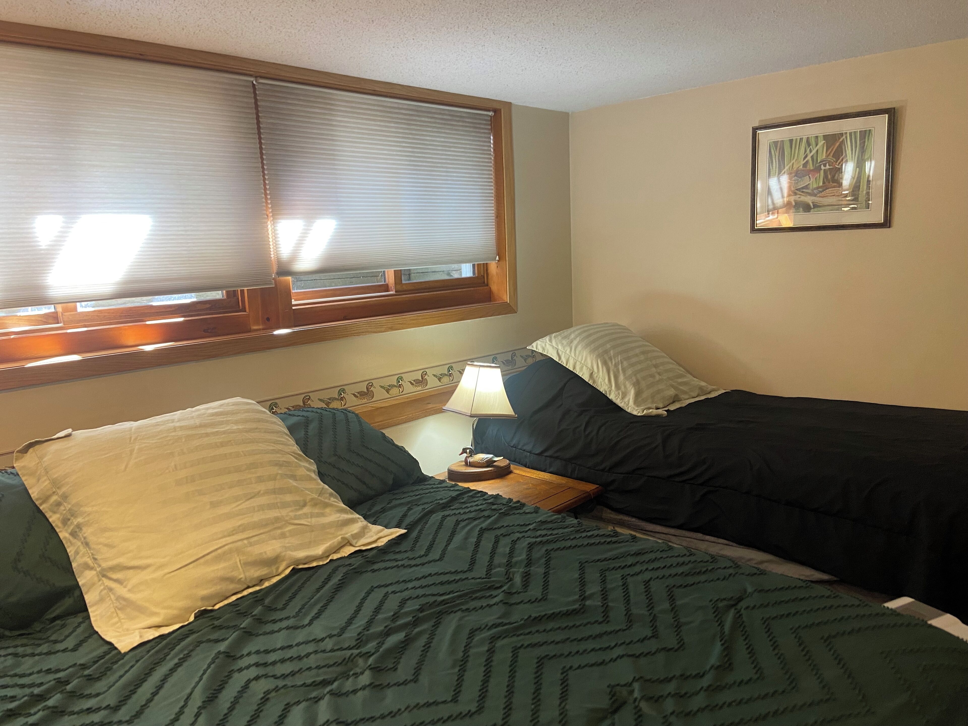 Un dormitorio con dos camas, una ventana con persianas, una mesita de noche con lámpara y un cuadro en la pared.