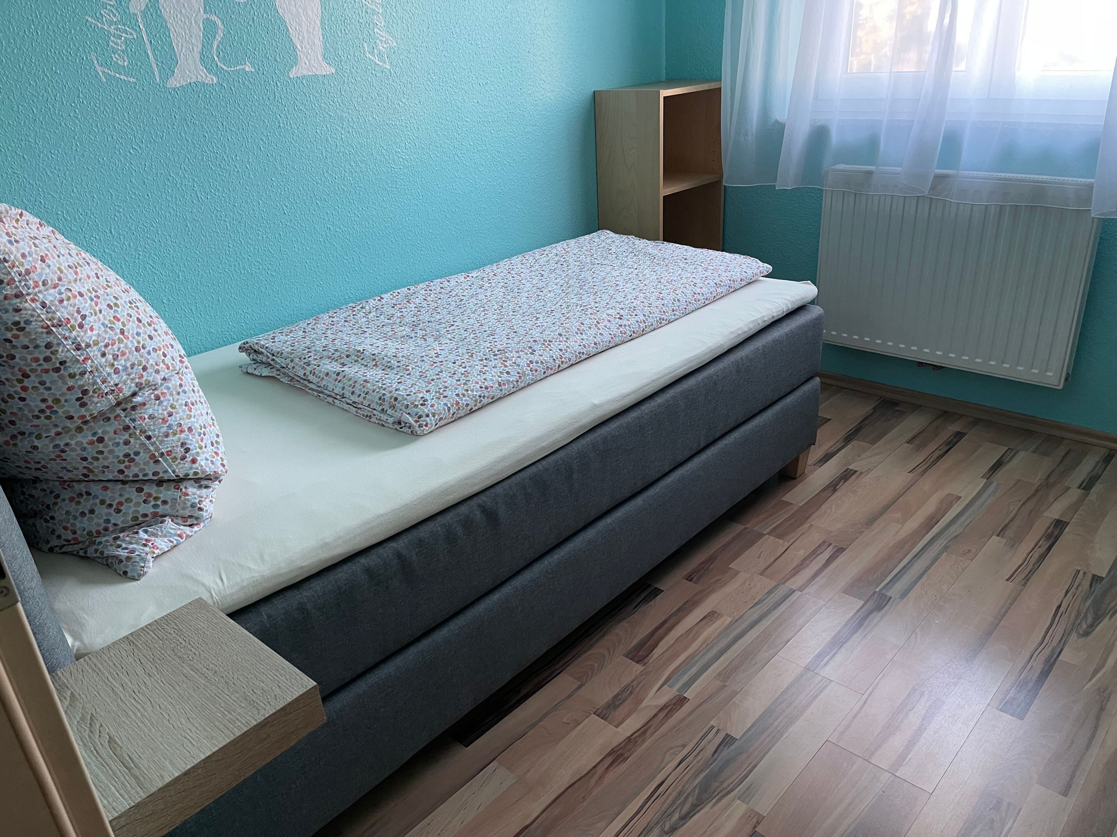 A small, neatly arranged room with a single bed, a wooden nightstand, a small wooden shelf, and a radiator.